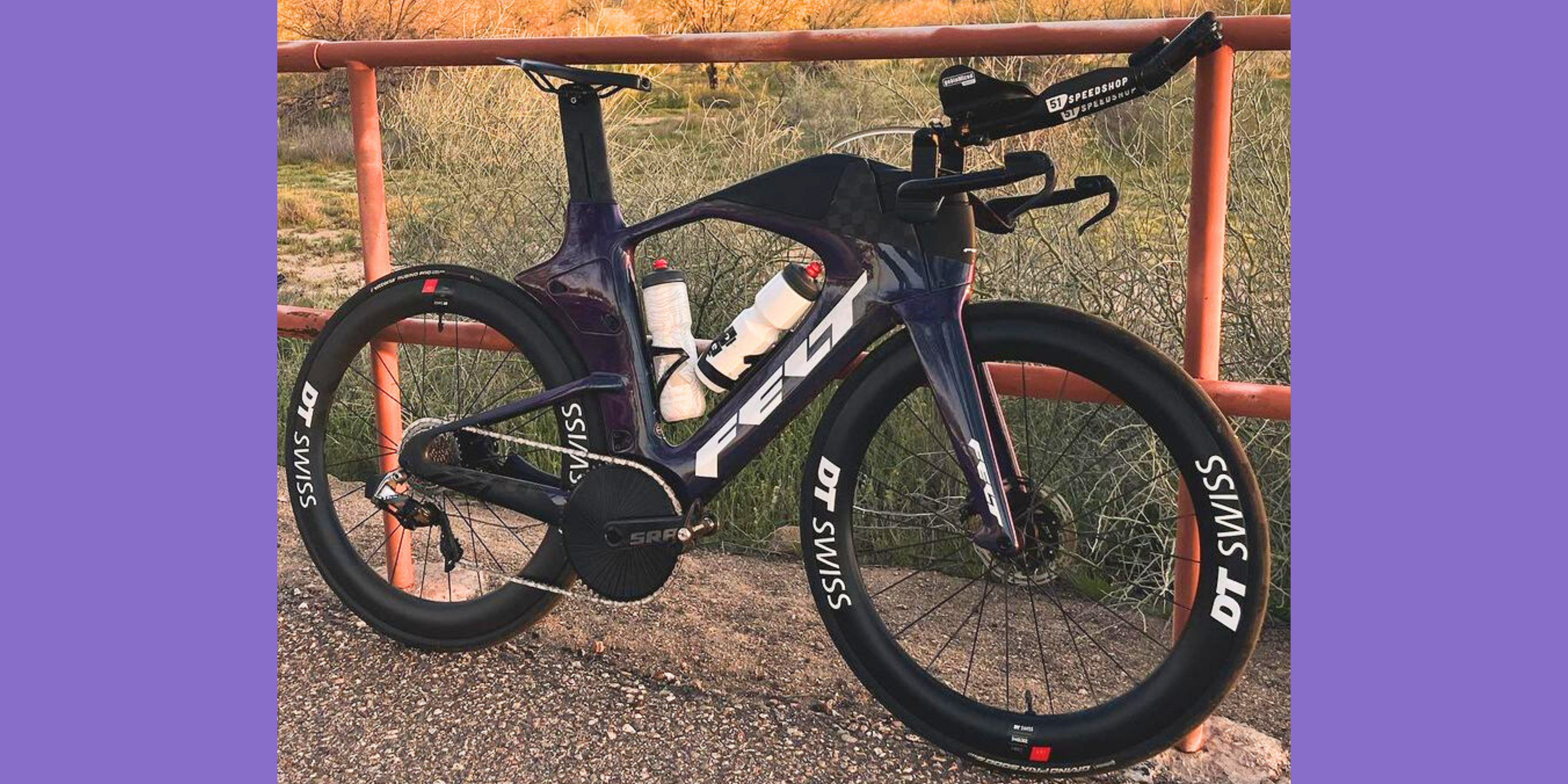  A close-up view of a high-end time trial bike branded "Felt" with DT Swiss wheels, two water bottles mounted in the frame, and a sleek aerodynamic design, parked against a rustic metal railing with a desert landscape in the background.